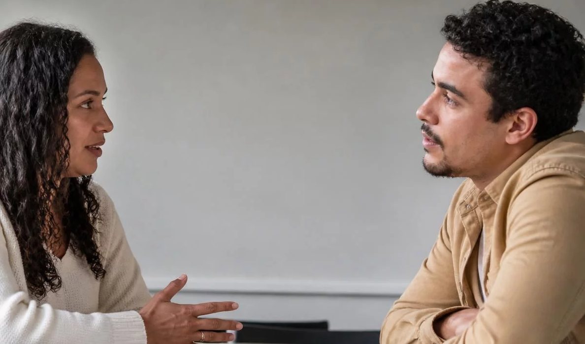 Três participantes conversam ao redor de uma mesa comprida numa sala clara de janelas altas